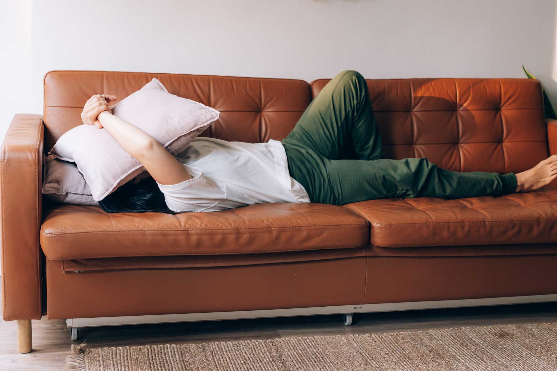 A depressed man covers his face with a pillow while laying on the couch.