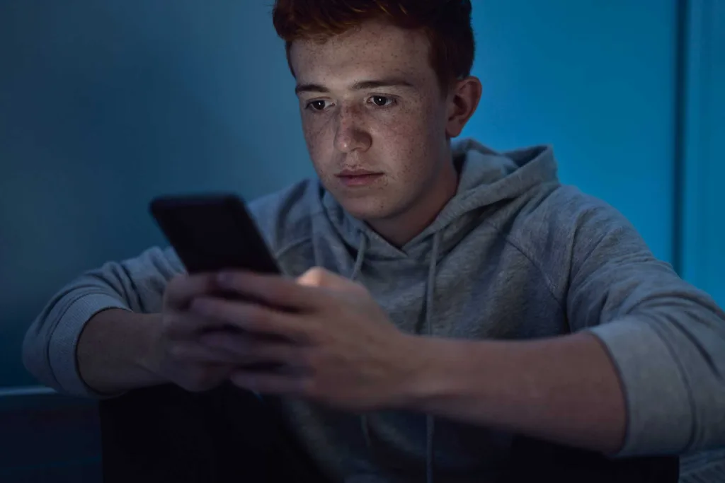 social media effects on youth illuistrated by a young man sitting alone in the dark as his face is illuminated by the glow of his smart phone